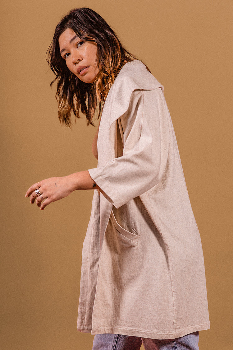 Model stanting up, as if walking, looking at the camera on a beije background. Blue jeans, light brown blouse, wearing a beije linen robe